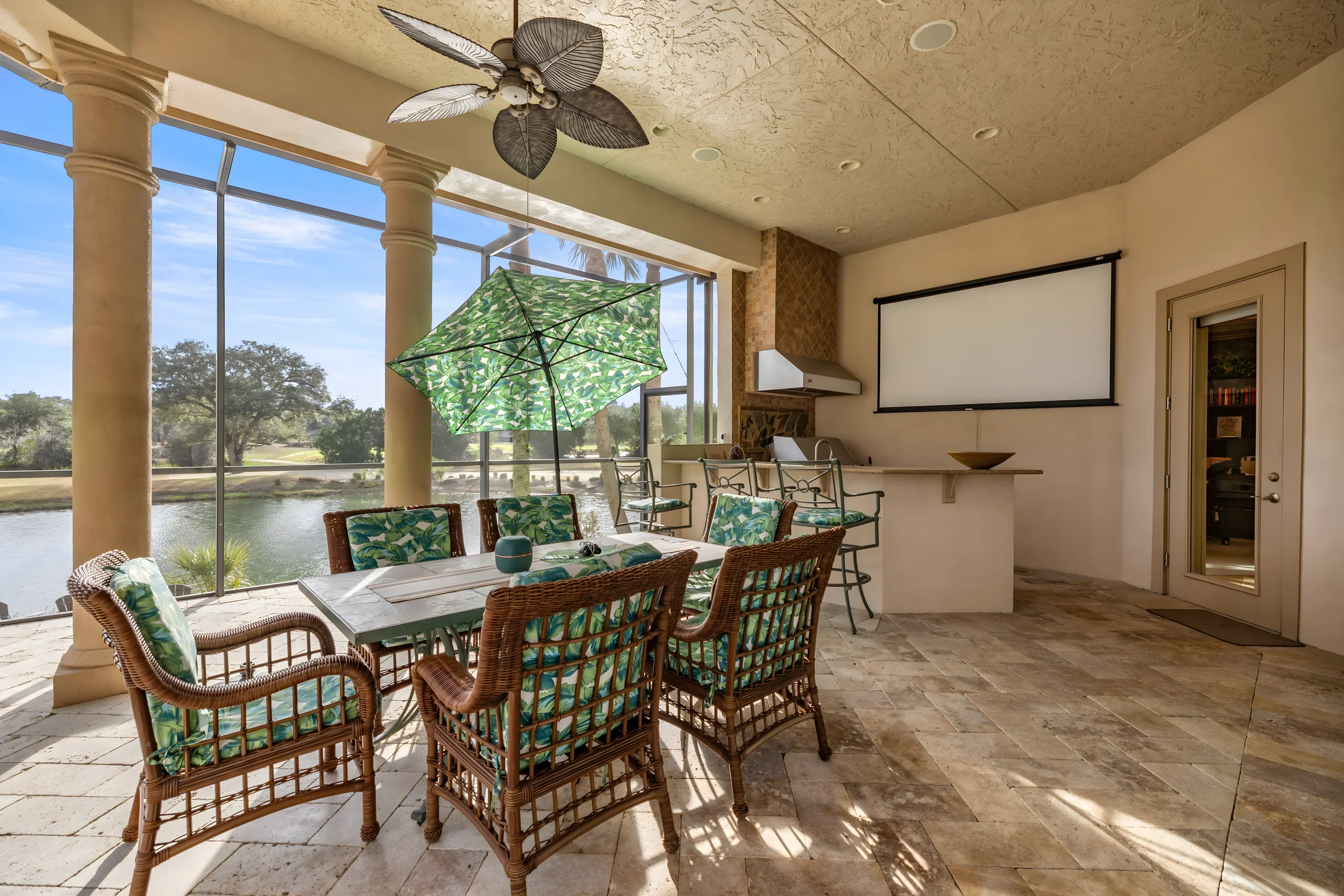 Covered lanai with a dining table, the outdoor kitchen and a projector screen, looking out to the pond