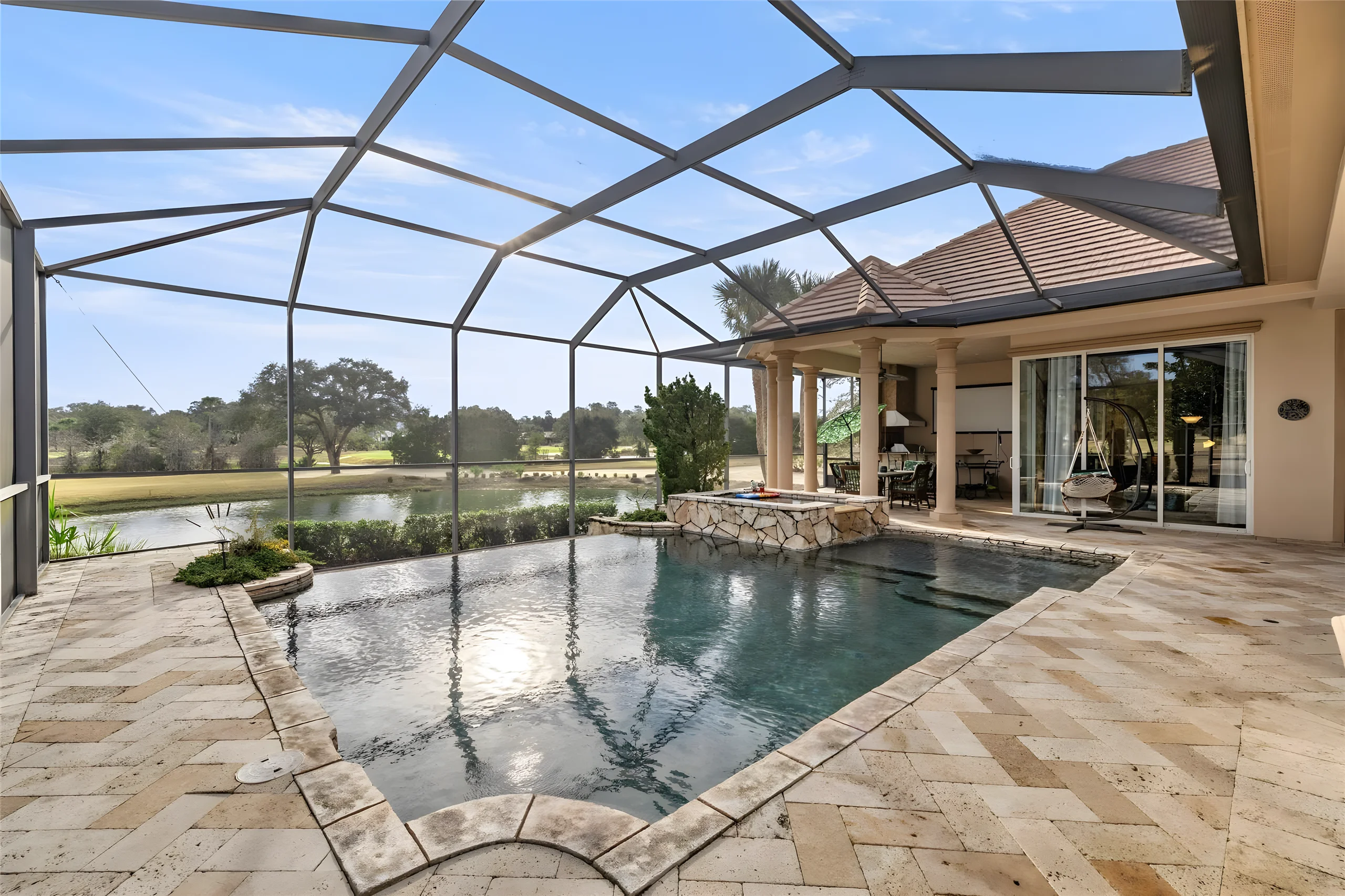 Pool deck in travertine with the pool and its raised stone spa, the pond and the golf course beyond the screen enclosure