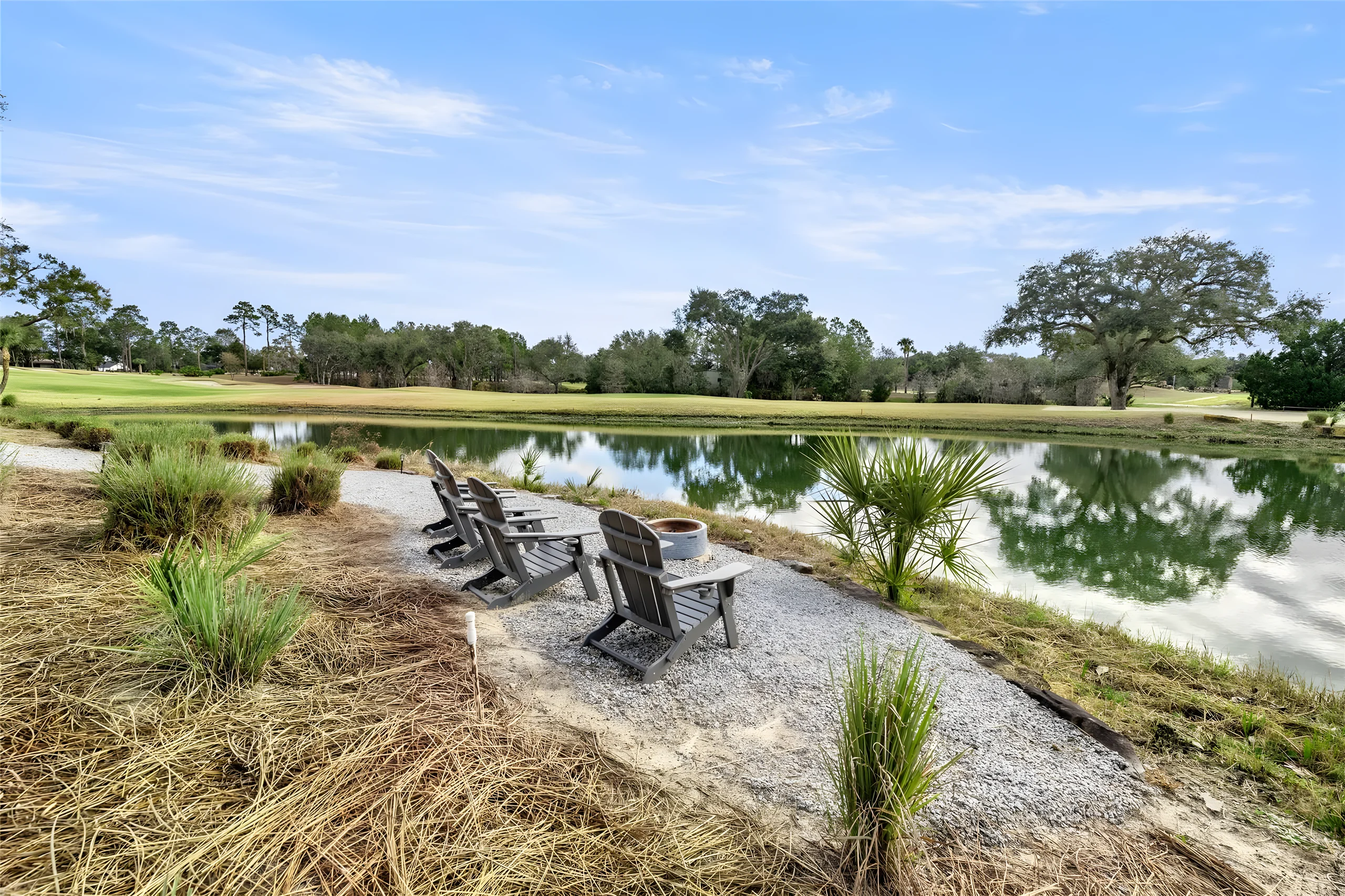 Adirondack chairs around a fire pit on a gravel sitting area at the edge of the pond, the golf course across the water